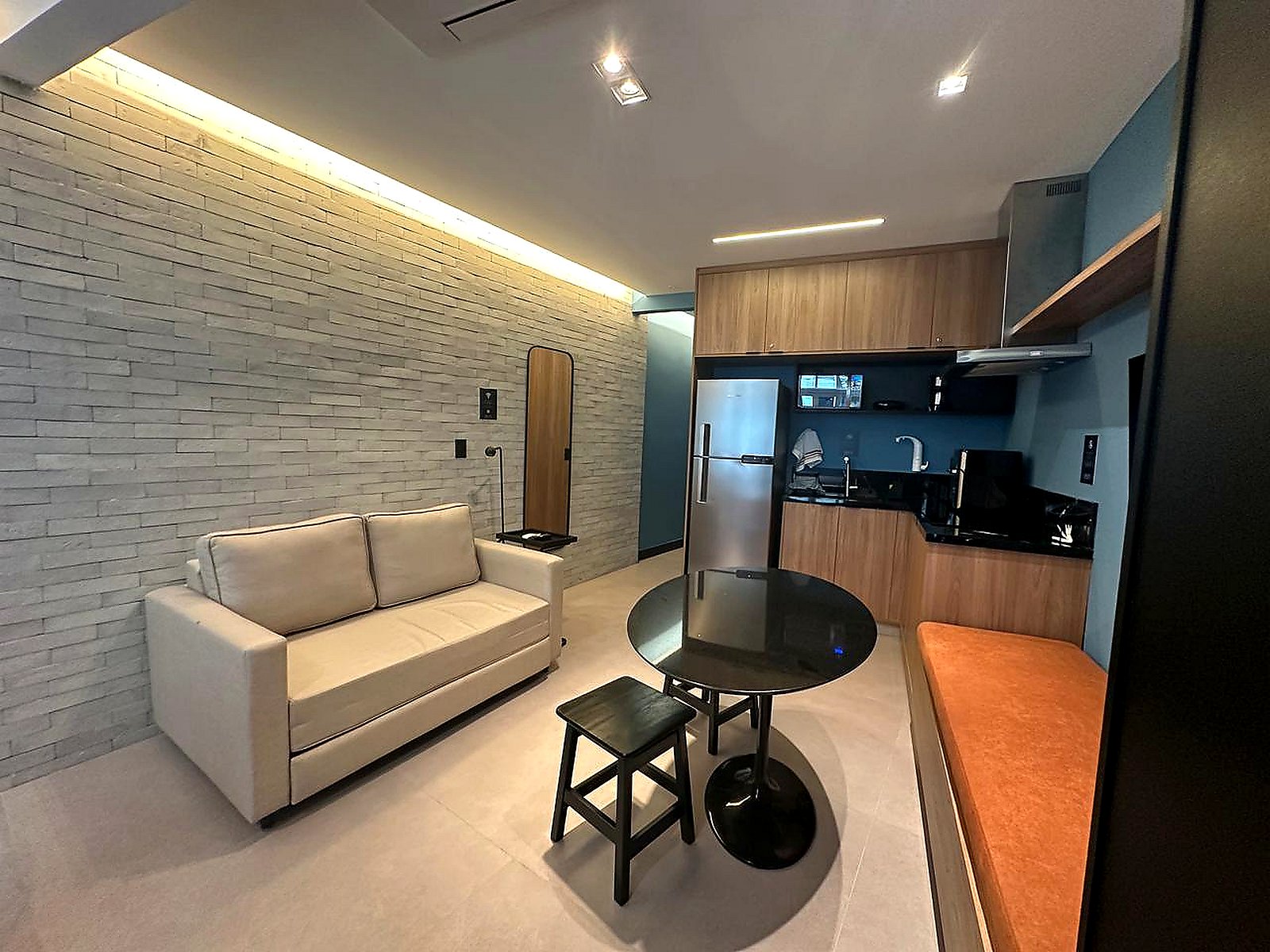 Sala e cozinha do Argos Safira, com sofá bege, mesa redonda preta e armários de madeira na cozinha, sem pessoas.